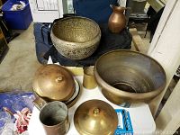View of multiple brass items including large bowls, covered bowls with lids, a small pitcher, and some cups on a white surface.