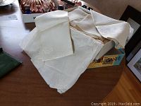 Box filled with various antique linens including napkins and cloths in off-white/cream color with floral patterns.
