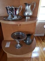 Display of six pieces: 5 pewter pieces on shelves with varying shapes including pitchers, sugar bowl, tray, and compote dish.