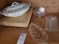 Photo showing covered floral Noritake serving dish with handles atop wooden cutting board, clear glass leaf-shaped dish, rectangular glass dish, and glass egg dish with decorative egg.