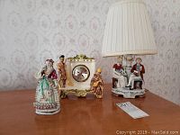 A wooden table displaying a porcelain lamp with seated couple base and white pleated lampshade, a porcelain clock with gold accents and small figurines, and a porcelain lady figurine dressed in 18th century style clothing.