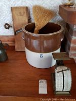 Kitchen counter with brown and cream crock marked 5, a small metal postage scale, flat wooden board, and hearth broom inside crock.