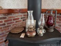 Three oil lamps on stove with brick backdrop: two traditional glass chimney and metal base lamps, and one red glass lantern oil lamp in metal frame.