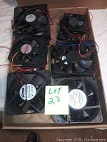 Group of 15 assorted mini electronic fans with black plastic frames and wires in a cardboard box, accompanied by a handwritten note 'LOT 23'.