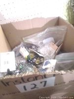 Open cardboard box containing several resealable plastic bags filled with various glass beads and marbles, including multi-colored and clear types.
