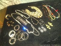 Photo shows a black background with an assortment of costume jewelry laid out including multiple necklaces, bracelets, bangles, and earrings.