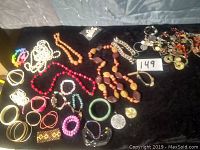 Overview of assortment of costume jewelry laid out on a dark cloth surface, shows variety of necklaces, bracelets, and a pile of smaller items including beads and earrings.