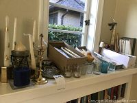 Display of assorted candles and candle holders arranged on a white windowsill, including wrapped taper candles and various glass holders.