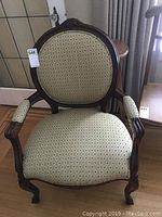Front view of antique wooden armchair with upholstered seat and backrest showing diamond patterned beige fabric and curved wooden arms and legs.