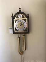 Front view of vintage wall clock with two brass weights and a pendulum hanging beneath. Wooden case with arched top and decorative finials.