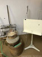 Overall photo showing multiple antique items including metal basket stand, woven basket, wooden spool, and white metal stand
