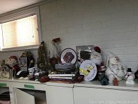 Wide view of shelf holding assorted holiday décor including plates, Santa figurines, fabric dolls, mugs, and small snowman figures