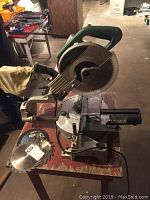 Chop saw mounted on wood stand with roller, showing entire unit and some blades on bench.