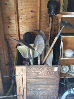 Photo showing assorted garden tools including shovels, spades, and a yard debris bag holder in a wooden container inside a shed.