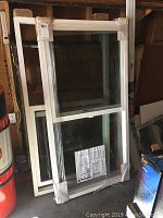 Two vertically standing white framed double glazed windows wrapped in plastic film, leaning against a wall in a garage or storage area. One window shows a specifications sticker.