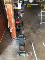 Full view of the blue hydraulic floor jack with black handle, metal wheels, and round lifting saddle on concrete floor.