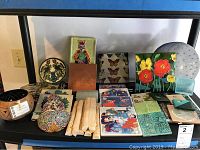 Full view of tile assortment displayed on shelf, showing a variety of tile sizes and artistic styles including floral, figurative, and geometric patterns.