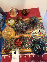 Photo showing eleven blown glass paper weights of varying sizes and colorful designs arranged on a patterned cloth.