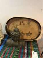 Photo showing bronze Buddha bust with detailed curls and closed eyes in front of rectangular wooden tray featuring Asian calligraphy and horsemen