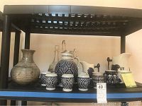 Photo showing the German ceramic decanter set with pewter lid, five matching cups, Sleepy Eye Pottery pitchers and cup, glass decanter, and pottery vase on black shelf.