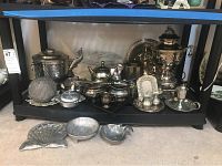 Wide view of multiple pewter, silverplate, and aluminum serving and decorative items on two shelves and the floor, arranged in groups including coffee urn, trays, pitchers, cream and sugar sets