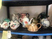 Shelf showing 9 tea pots of various shapes, colors and decorations, mostly porcelain or ceramic with floral and bird motifs, some chipped.