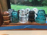 Nine glass and ceramic insulators on windowsill in natural light showing variety of colors and shapes including clear, seafoam blue, and brown items.