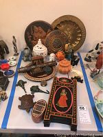 A large assortment of Asian decorative items including metal plates, carved wooden figurines, elephant incense holder, painted small furniture, and ceramic jars.