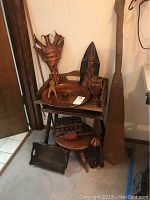 Two tier wooden table with various wooden and leather art pieces displayed: wooden trays, goblets, carved box, leather face sculptures, wooden stool underneath and small carved figures.