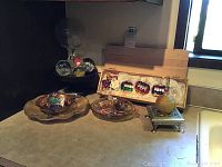 Photo showing various items including wooden pedestal, Asian plate, glass paperweights, and Kurt S Adler train ornaments in wooden crate on countertop.