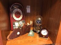Shelf display of five clocks including a large wooden wall clock with red face, a natural edge wood plaque clock, a granite pyramid clock, a Bulova clock in glass dome, and a small polished Seiko clock.