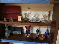 Full view of two wooden shelves showing a mix of glass perfume bottles, a red and white vase, a blue and white bottle, mirror plate, two long decorative boxes, and a tall brass vase.