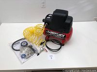 Full view of Husky 2 gallon air compressor with attached yellow coiled hose. Accessories in plastic bag nearby.