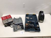 Full lot overview showing battery charger, heat gun kit with blue case, and rotary tool kit with grey case