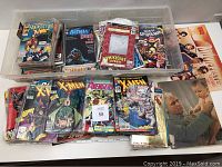 Top-down view of assorted comic books and memorabilia in plastic bin showing covers of Web of Spider-Man, Batman, Dark Knight, Death in the Family, Fantastic Four, X-Men, Avengers