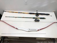 Two fishing rods and a white Bear bow with red limbs laid on a table