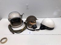 Three helmets on a table: white pilot helmet at left, olive drab steel helmet with gas mask center, white open-face helmet right