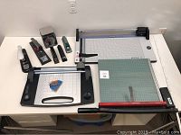 Wide view of the lot showing three paper cutters, five staplers, and one postal mail scale arranged on a white surface.