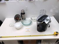 Full view of Keurig coffee maker, Pyrex bowls and plates, Thermos flask, metal strainer, and jug collection on white table.