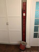 Straight-on view showing tall red Safeco fire blanket case with white lettering, placed against wall next to white cabinets, accompanied by a red fire bucket at base.