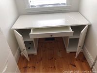 Frontal view of white IKEA desk showing open drawer and cabinet doors, wooden floor beneath.