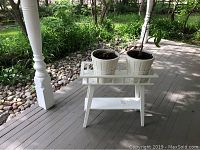 Plant stand with two white pots and plants on porch floor with decorative post and stones in background.
