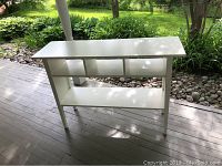 Front view of white IKEA Lyckhem sofa table showing top surface, three open compartments, and bottom shelf on a porch wooden floor with green outdoor background.