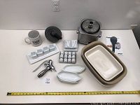 Photo showing the full lot including the Cuisinart rice cooker, casserole dishes, wine opener, corn holders, salt and pepper shakers, coffee mug, mortar and pestle, and serving dishes laid out on a table with measuring tape for scale.