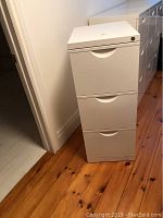 White 3-drawer filing cabinet standing against wall on wooden floor, side angle view showing drawer handles and lock on top drawer.