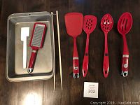 Red KitchenAir utensils, two metal trays, white plastic spatula and two wooden chopsticks laid out on dark table.