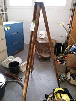 Full view of vintage wooden tripod standing upright on a concrete floor surrounded by other workshop items.