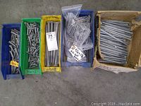 Photo showing multiple containers with metal bolts of various lengths and diameters. Includes green, yellow, blue plastic containers and a cardboard box. Labels indicate bolt sizes and types.