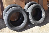 Four black automotive tires arranged outdoors on ground by wooden wall, used condition, sunlight visible.