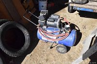 Photo showing full side view of the blue Campbell Hausfeld compressor on wheels with hose coiled around it next to a tire